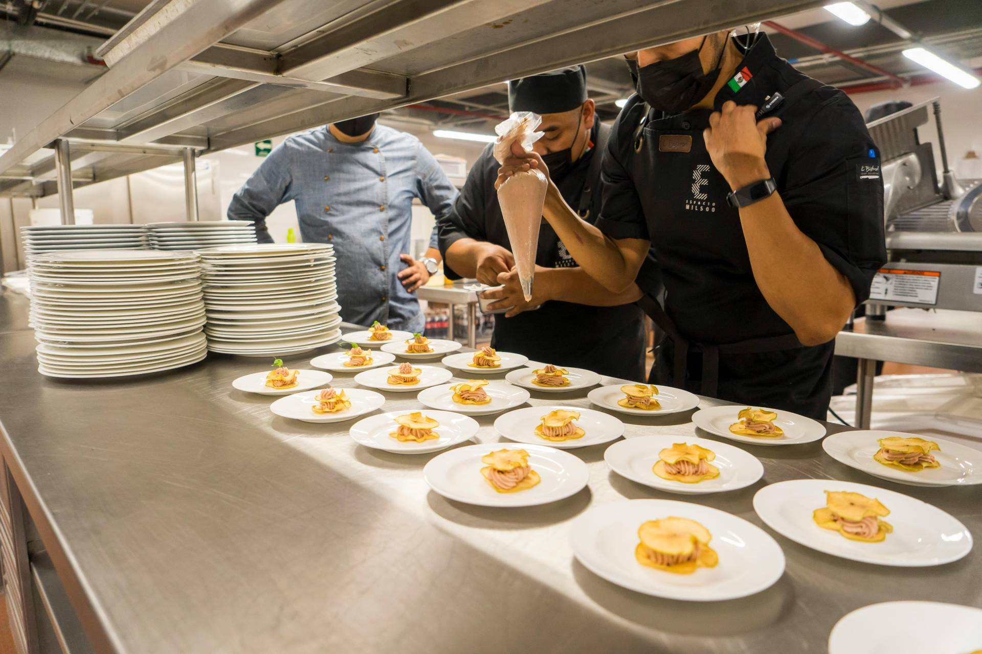 Chefs in a professional kitchen preparing multiple plates of finely arranged appetizers. One chef uses a piping bag to add a finishing touch to the dishes while another chef observes. Stacks of clean plates are visible beside them, emphasizing attention to detail and cleanliness in a high-end culinary setting.