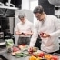 Two chefs working in a professional kitchen. One chef is writing on a clipboard, while the other is holding an onion and preparing vegetables. The counter is filled with fresh ingredients including peppers, tomatoes, and herbs.