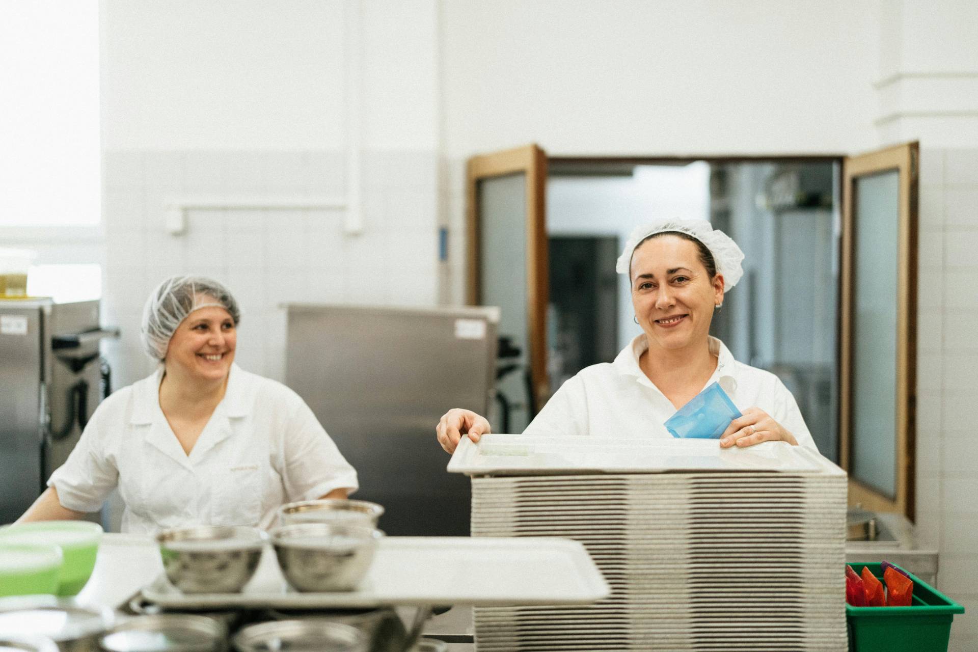 Two women in a kitchen wearing white uniforms and hairnets. One is smiling near a stack of trays, while the other holds onto the edge of the trays with a pleasant expression. The kitchen environment is clean and organized, emphasizing hygiene and food safety practices.