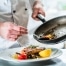 Chef plating a dish with seared salmon, roasted vegetables, and garnish, carefully spooning sauce from a skillet onto the plate in a professional kitchen setting.