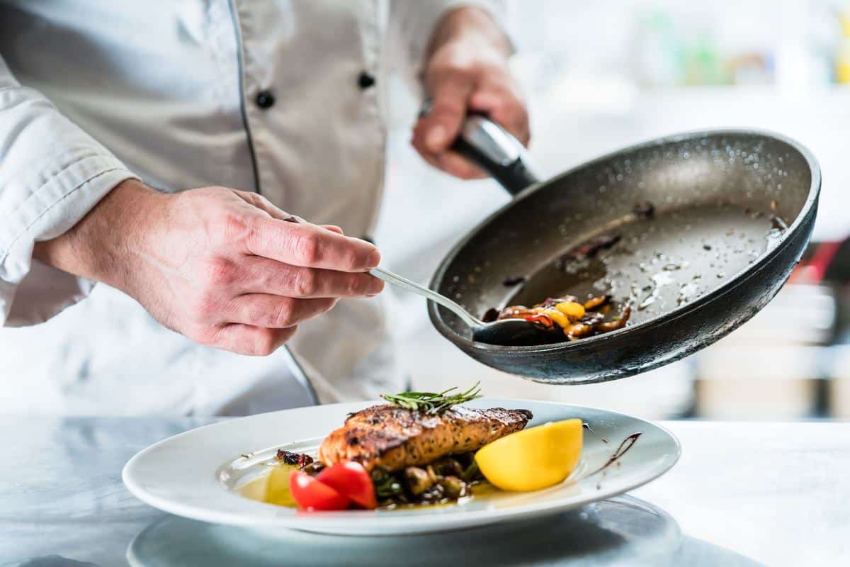 Chef plating a dish with seared salmon, roasted vegetables, and garnish, carefully spooning sauce from a skillet onto the plate in a professional kitchen setting.